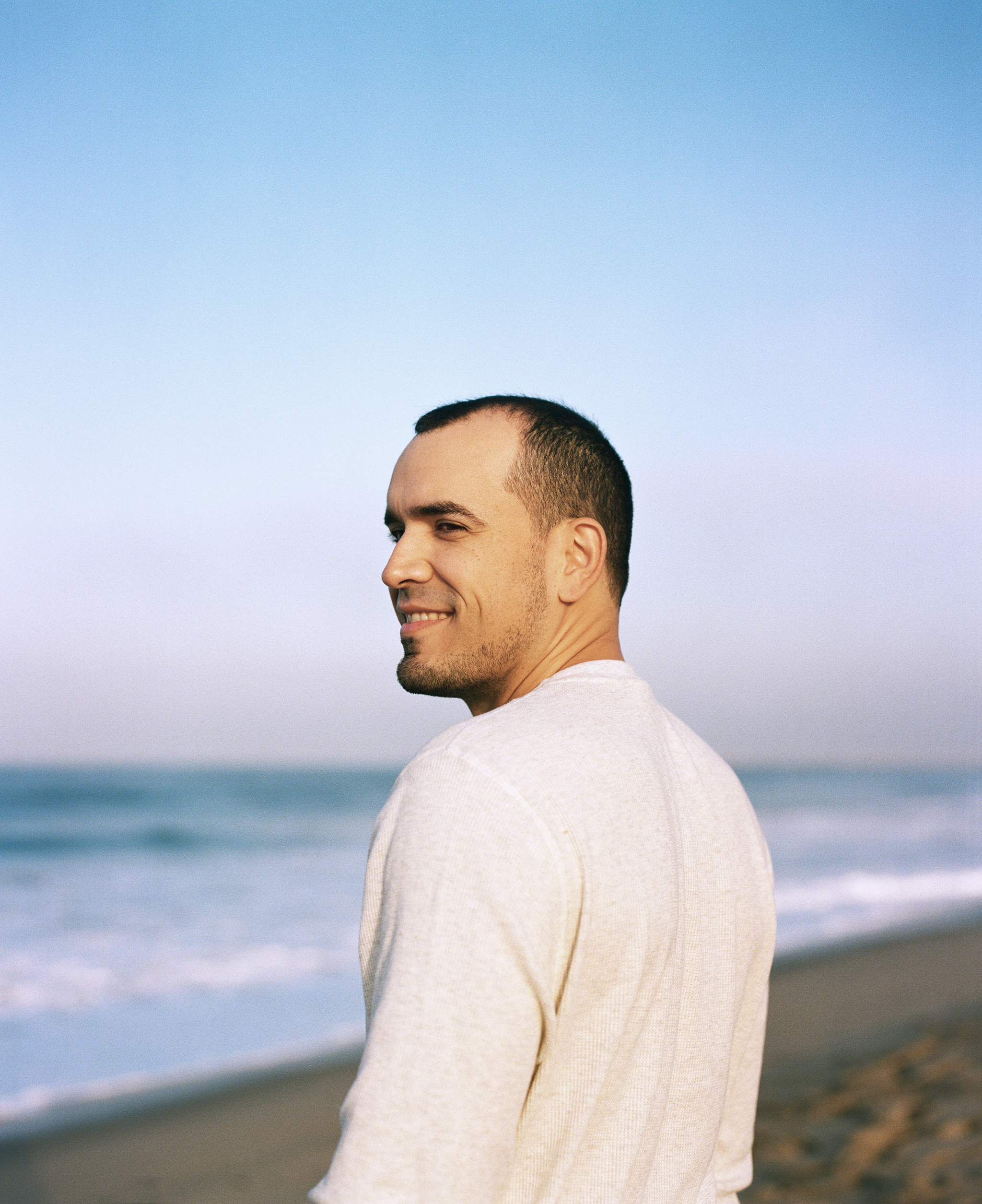 A man stands in front of the ocean, smiling. He is looking over his shoulder and has a white shirt on